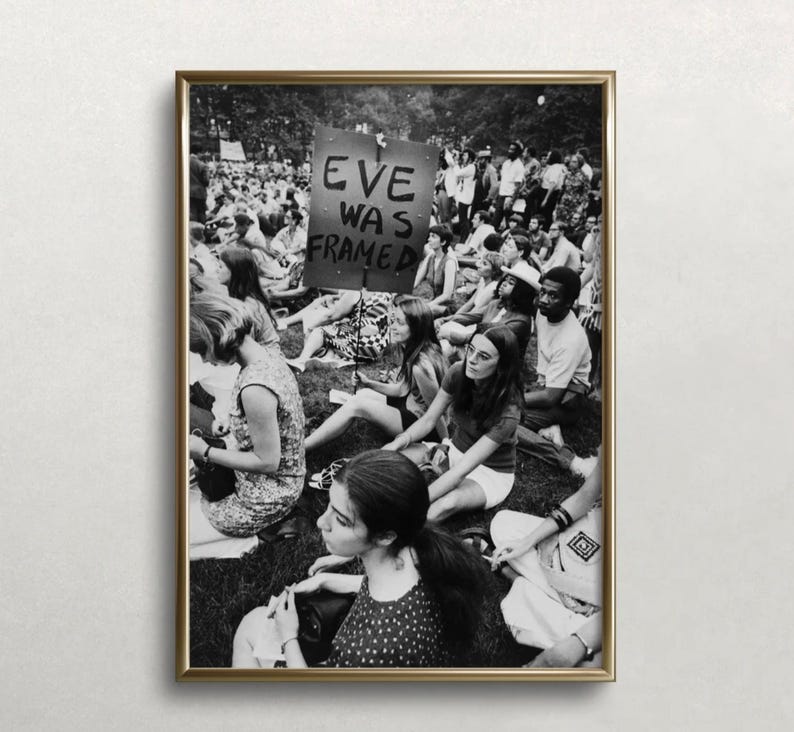 May include: Black and white photograph of a group of people sitting on the grass. A sign reads "Eve Was Framed".