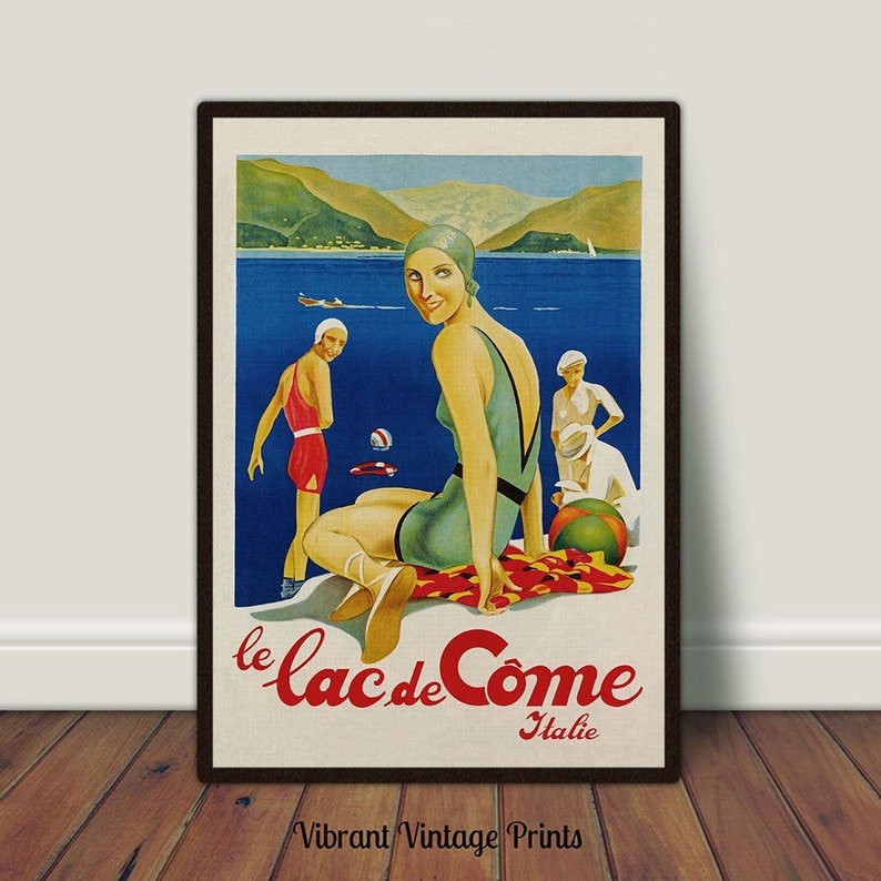 May include: Vintage travel poster print featuring a woman in a green swimsuit and cap, seated near a lake. The poster has the text "le Lac de C&ocirc;me Italie" and depicts other figures on a beach with mountains in the background. The print is framed.