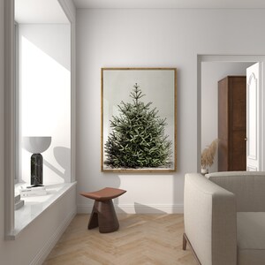 May include: A framed botanical print of a green evergreen tree. The artwork is displayed in a bright, modern room with a wooden frame. The room also features a wooden stool, a marble lamp, and a neutral-toned sofa.