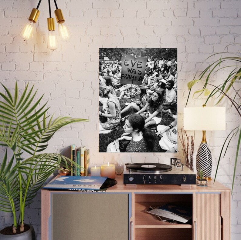 Puede incluir: Fotograf&iacute;a en blanco y negro de un grupo de personas sentadas en la hierba. Un cartel en el centro de la imagen dice "Eve Was Framed".