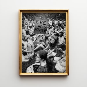 May include: A black and white framed photograph depicting a crowd of people sitting on grass at an outdoor gathering. A prominent sign held in the center displays the text EVE WAS FRAMED in bold, hand-painted black capital letters on a light background.