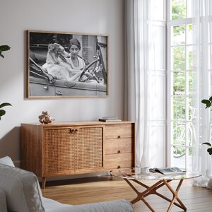 May include: Black and white framed photograph of a woman and a dog in a vintage car. The photograph is displayed above a wooden cabinet with a woven door. A glass coffee table and white curtains are also visible.