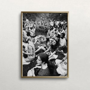 Puede incluir: Fotograf&iacute;a en blanco y negro de un grupo de personas sentadas en la hierba. Un cartel en el centro de la imagen dice "Eve Was Framed".