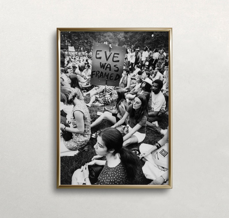 Puede incluir: Fotograf&iacute;a en blanco y negro de un grupo de personas sentadas en la hierba. Un cartel en el centro de la imagen dice "Eve Was Framed".