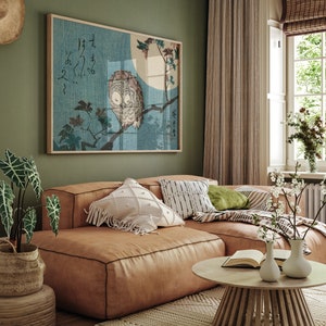 May include: A brown leather sectional sofa with white and black patterned pillows, a green pillow, and a throw blanket. A wooden coffee table with a book and two white vases with flowers sits in front of the sofa. A framed print of an owl perched on a branch with Japanese characters hangs on the wall behind the sofa.