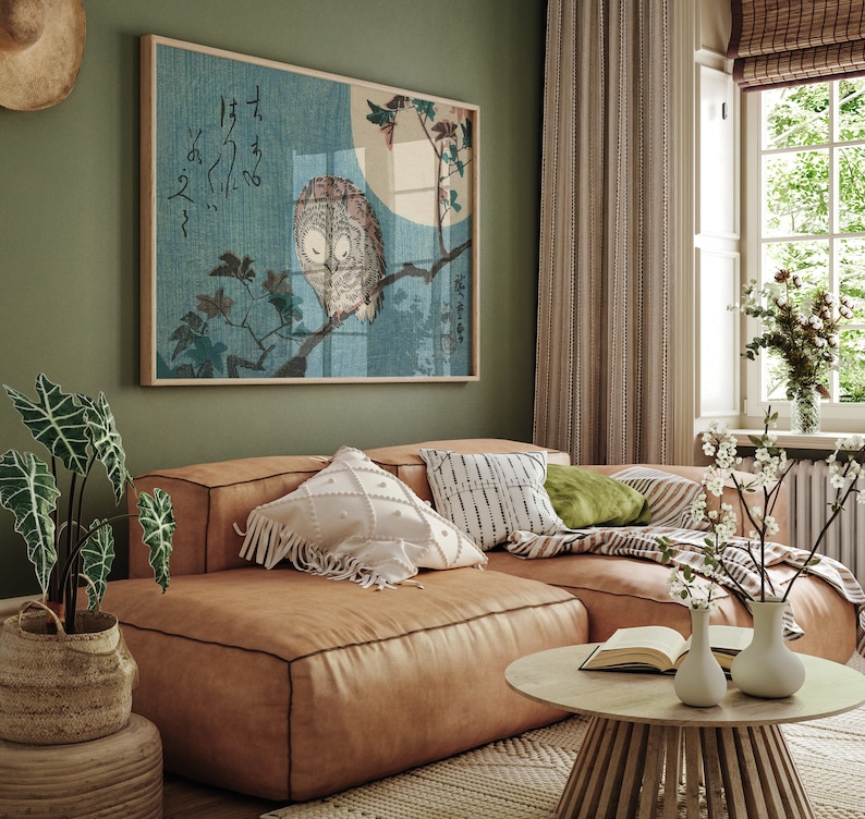 May include: A brown leather sectional sofa with white and black patterned pillows, a green pillow, and a throw blanket. A wooden coffee table with a book and two white vases with flowers sits in front of the sofa. A framed print of an owl perched on a branch with Japanese characters hangs on the wall behind the sofa.