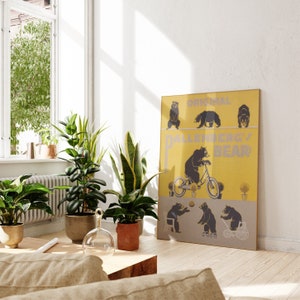 Op de afbeelding: Een ingelijste poster met een gele en grijze achtergrond toont illustraties van beren. De tekst "ORIGINAL BALLENBERG'S BEAR" is zichtbaar. De poster leunt tegen een witte muur, in de buurt van potplanten en een houten salontafel.