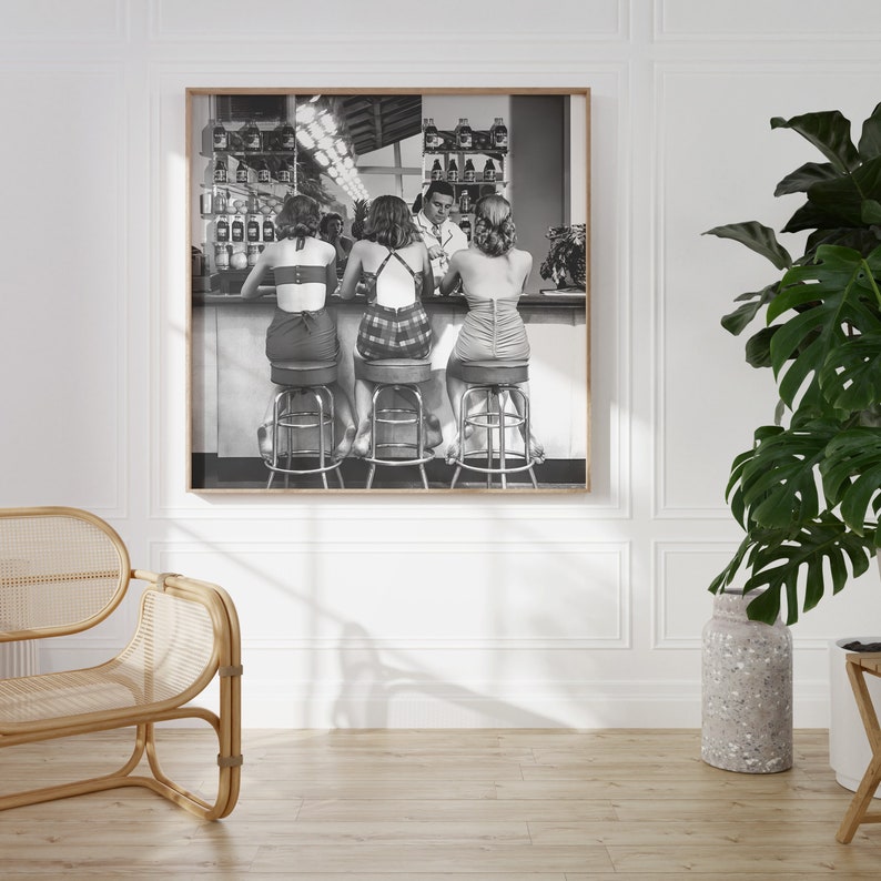 Op de afbeelding: Zwart-witfoto van drie vrouwen die op barstoelen aan een bar zitten. De vrouwen dragen kleding in de stijl van de jaren 1950 en kijken met hun rug naar de camera. Een man staat achter de bar en kijkt naar de vrouwen.