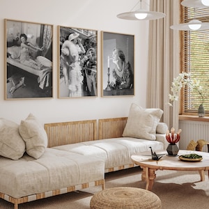 May include: Three black and white framed photographs of women in vintage clothing. The photos are hanging on a white wall above a light brown sofa with white cushions. The sofa is in a living room with a wooden coffee table and a woven rug.