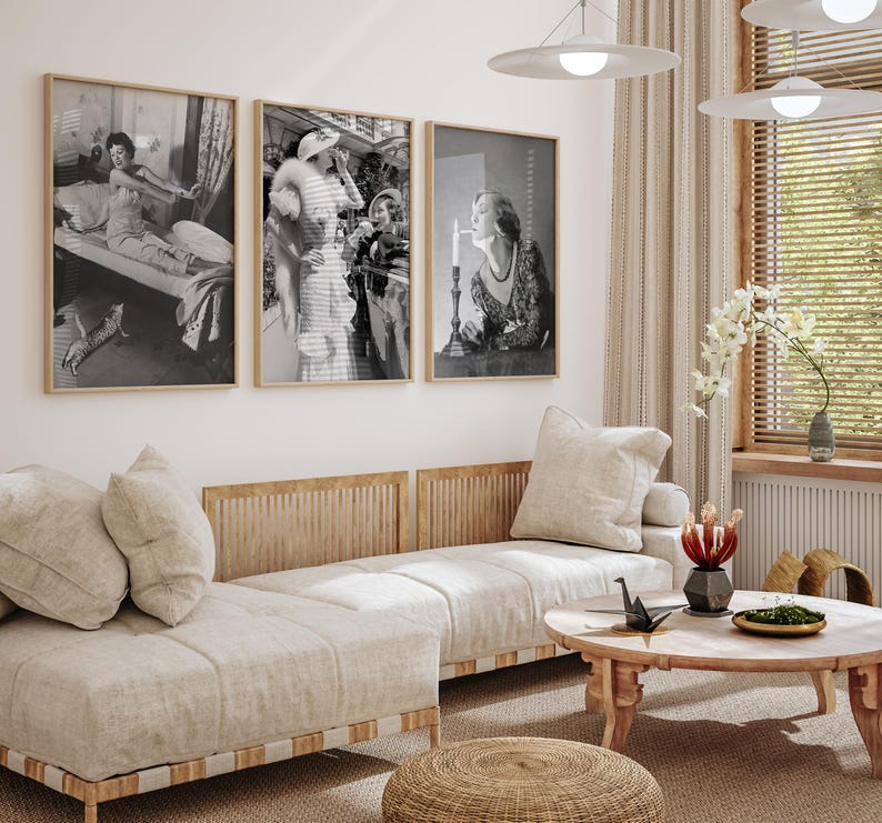 May include: Three black and white framed photographs of women in vintage clothing. The photos are hanging on a white wall above a light brown sofa with white cushions. The sofa is in a living room with a wooden coffee table and a woven rug.