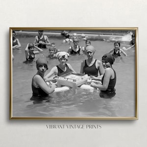 May include: Black and white vintage photograph of a group of women in swimsuits playing a game in a swimming pool. The women are wearing swim caps and are sitting on a floating platform. The game appears to be a card game.