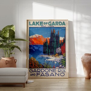 May include: A framed vintage travel poster for Lake Garda, Italy, featuring a scenic landscape with mountains, trees, and a lake. The poster has text that reads "Lake of Garda Golf Links" and "Gardone Riv. and Fasano". The poster is displayed in a room with a plant and a footstool.