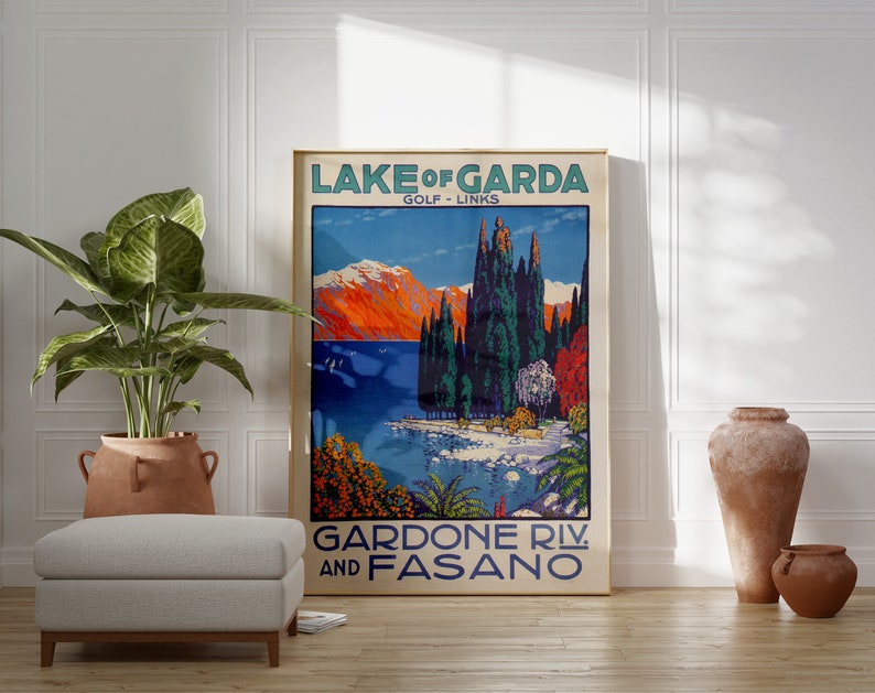 May include: A framed vintage travel poster for Lake Garda, Italy, featuring a scenic landscape with mountains, trees, and a lake. The poster has text that reads "Lake of Garda Golf Links" and "Gardone Riv. and Fasano". The poster is displayed in a room with a plant and a footstool.