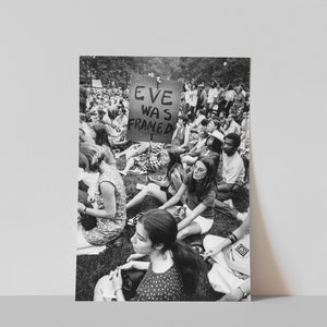 Puede incluir: Una fotograf&iacute;a en blanco y negro de un grupo de personas sentadas en la hierba. Un cartel dice "Eve Was Framed".