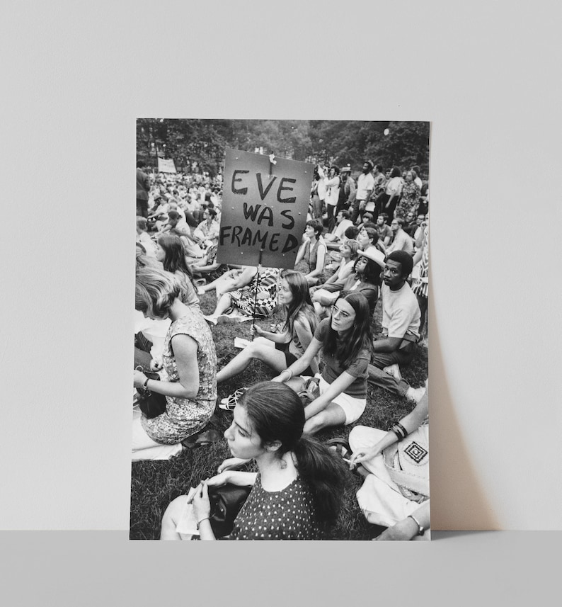 Puede incluir: Una fotograf&iacute;a en blanco y negro de un grupo de personas sentadas en la hierba. Un cartel dice "Eve Was Framed".