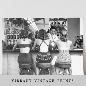Op de afbeelding: Zwart-witfoto van drie vrouwen die aan een bar zitten in badpakken. De vrouwen kijken naar een man in een wit overhemd die achter de bar staat. De bar heeft een plank met flessen erop. De vrouwen zitten op krukken.