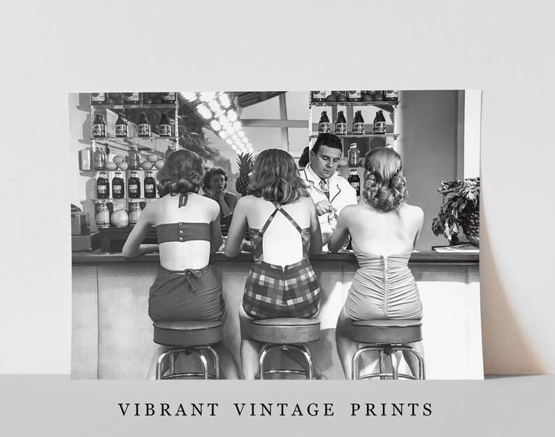 Op de afbeelding: Zwart-witfoto van drie vrouwen die aan een bar zitten in badpakken. De vrouwen kijken naar een man in een wit overhemd die achter de bar staat. De bar heeft een plank met flessen erop. De vrouwen zitten op krukken.