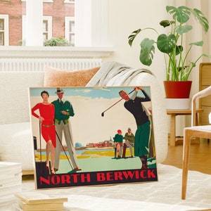 May include: A framed vintage golf poster with the text "NORTH BERWICK" in red. The poster features illustrations of golfers and figures in vintage attire. The poster is displayed in a living room setting with a plant and a couch.