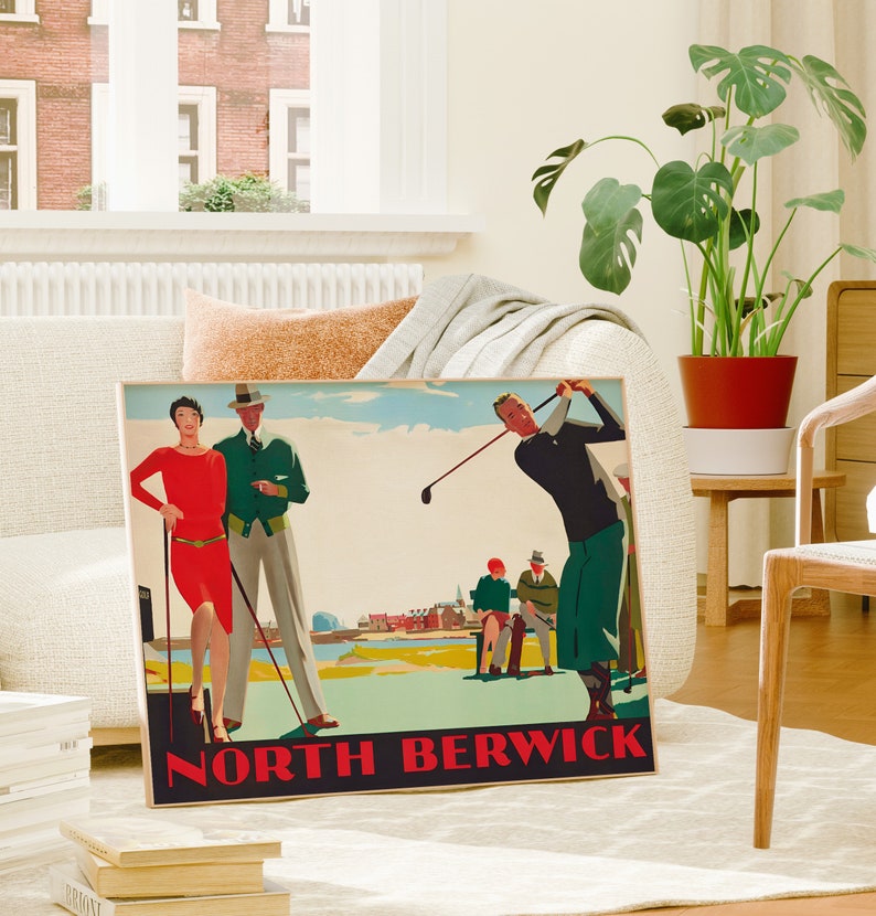 May include: A framed vintage golf poster with the text "NORTH BERWICK" in red. The poster features illustrations of golfers and figures in vintage attire. The poster is displayed in a living room setting with a plant and a couch.