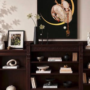 Op de afbeelding: Een donkerhouten boekenplank met meerdere planken gevuld met boeken, decoratieve schalen en sokkels. Boven de plank hangt een ingelijste kunstdruk met een figuur in een kledingstuk met patronen, zittend in een gouden halve maan op zwart.