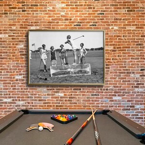 May include: A black and white photograph of four women playing golf. The women are wearing dresses and hats. The photograph is framed in a silver frame and hanging on a brick wall. A pool table is in the foreground.