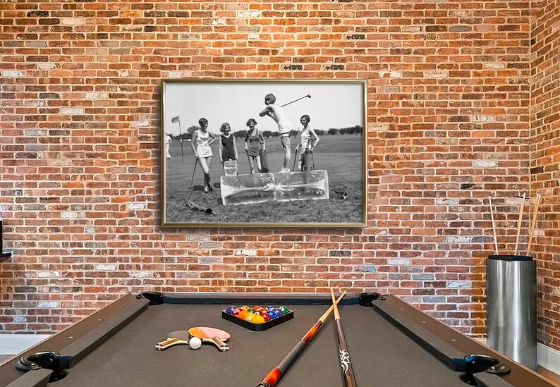 May include: A black and white photograph of four women playing golf. The women are wearing dresses and hats. The photograph is framed in a silver frame and hanging on a brick wall. A pool table is in the foreground.