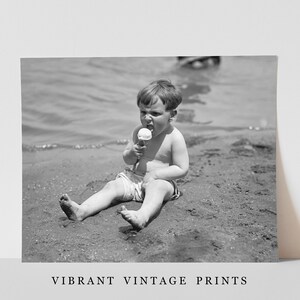May include: A young boy sits on a sandy beach, wearing a white bathing suit and eating an ice cream cone.
