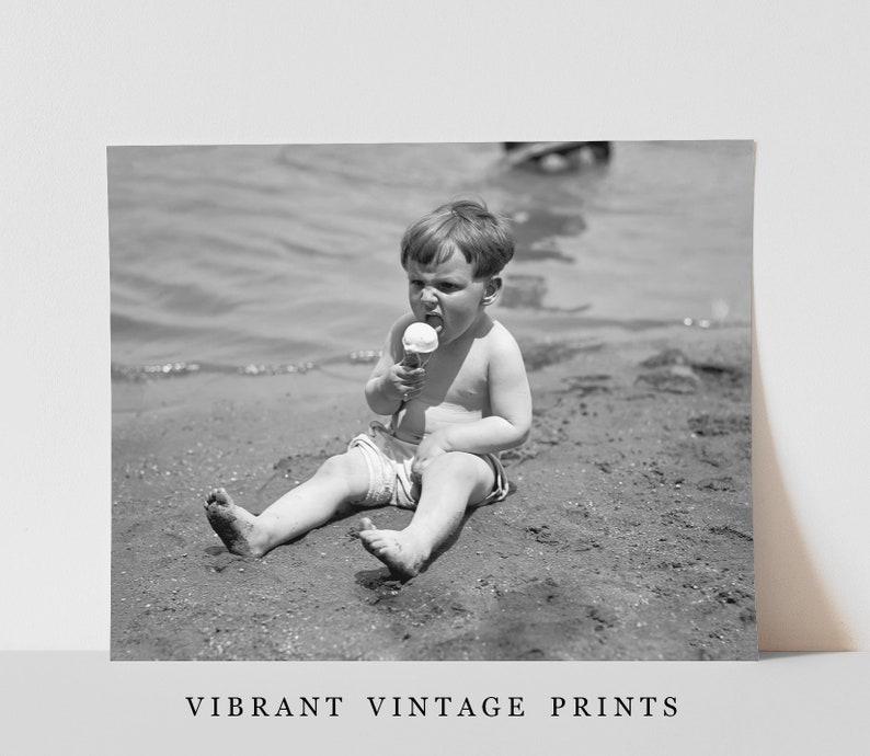 May include: A young boy sits on a sandy beach, wearing a white bathing suit and eating an ice cream cone.