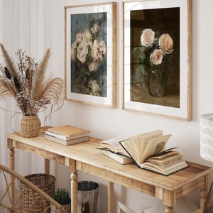 May include: Two framed floral paintings with a light brown wooden frame. The paintings feature bouquets of pink roses in a vase. The paintings are hanging on a white wall above a wooden console table with books and a vase of dried flowers.