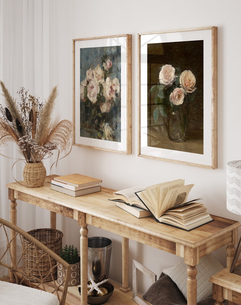 May include: Two framed floral paintings with a light brown wooden frame. The paintings feature bouquets of pink roses in a vase. The paintings are hanging on a white wall above a wooden console table with books and a vase of dried flowers.