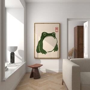 May include: A framed print of a green frog with a white background. The frog is facing forward with its mouth closed. The print has a red square with the Japanese character "開" in the top right corner.