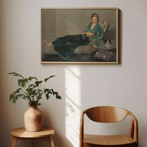 May include: A framed painting of a woman in a green dress sitting on a couch and reading a book. The painting is in a light-colored frame and is hanging on a white wall.
