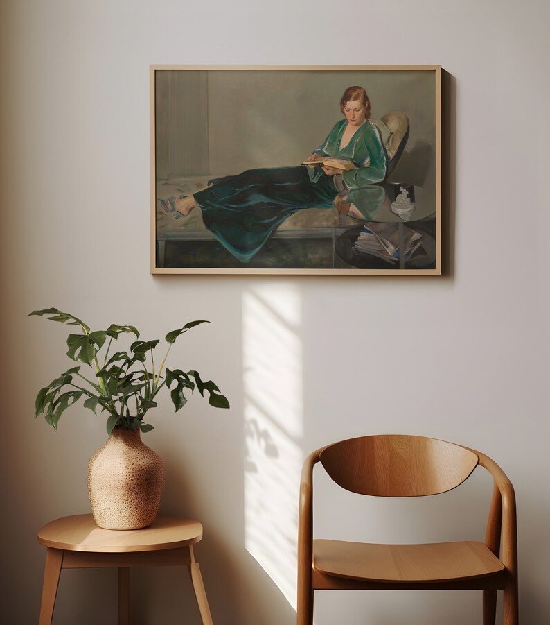May include: A framed painting of a woman in a green dress sitting on a couch and reading a book. The painting is in a light-colored frame and is hanging on a white wall.