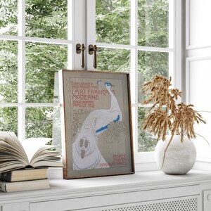 Op de afbeelding: Een ingelijste vintage kunstprint met een witte pauw en blauwe accenten. De poster, met Franse tekst, wordt tentoongesteld op een witte vensterbank. Er is een open boek, een vaas met gedroogde planten en een raam op de achtergrond.