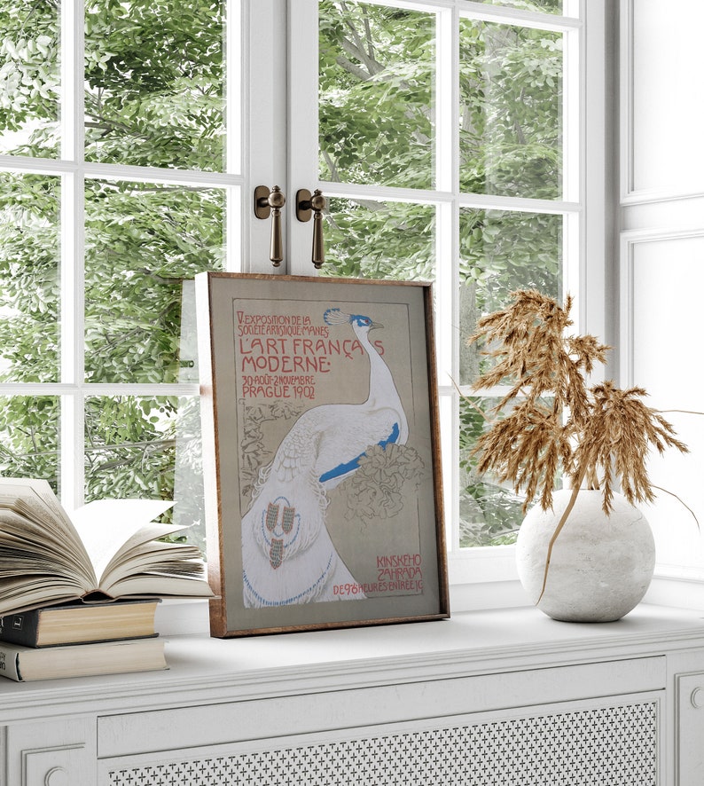 Op de afbeelding: Een ingelijste vintage kunstprint met een witte pauw en blauwe accenten. De poster, met Franse tekst, wordt tentoongesteld op een witte vensterbank. Er is een open boek, een vaas met gedroogde planten en een raam op de achtergrond.