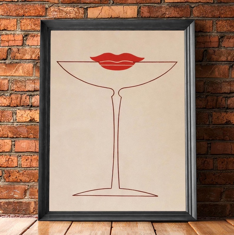 May include: A framed wall art print featuring a minimalist line drawing of a martini glass in dark red on a beige background, with a pair of solid red lips positioned directly above the rim of the glass, enclosed in a simple black frame.