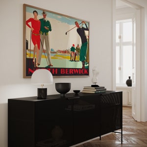 May include: A framed vintage golf poster with the text "North Berwick" hangs above a black cabinet. The poster depicts golfers in a colorful, stylized illustration. A white lamp, black bowl, and books sit on the cabinet.
