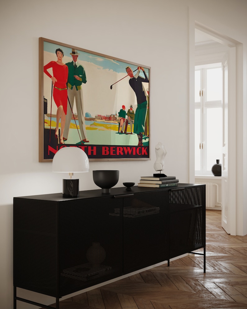 May include: A framed vintage golf poster with the text "North Berwick" hangs above a black cabinet. The poster depicts golfers in a colorful, stylized illustration. A white lamp, black bowl, and books sit on the cabinet.