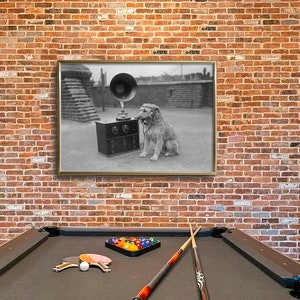 May include: A framed black and white photograph of a dog listening to a vintage gramophone. The dog is wearing headphones and sitting next to the gramophone. The photograph is hanging on a brick wall above a pool table.