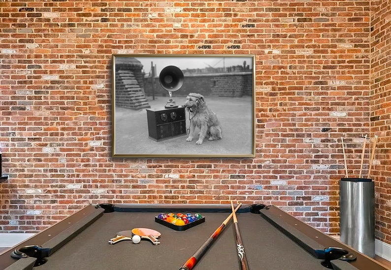 May include: A framed black and white photograph of a dog listening to a vintage gramophone. The dog is wearing headphones and sitting next to the gramophone. The photograph is hanging on a brick wall above a pool table.