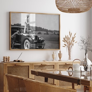 Op de afbeelding: Een zwart-witfoto van een man die op de treeplank van een vintage auto staat en een golfclub vasthoudt. De auto staat geparkeerd op een grasveld. De foto is ingelijst en hangt aan een witte muur boven een houten kast met rieten deuren.