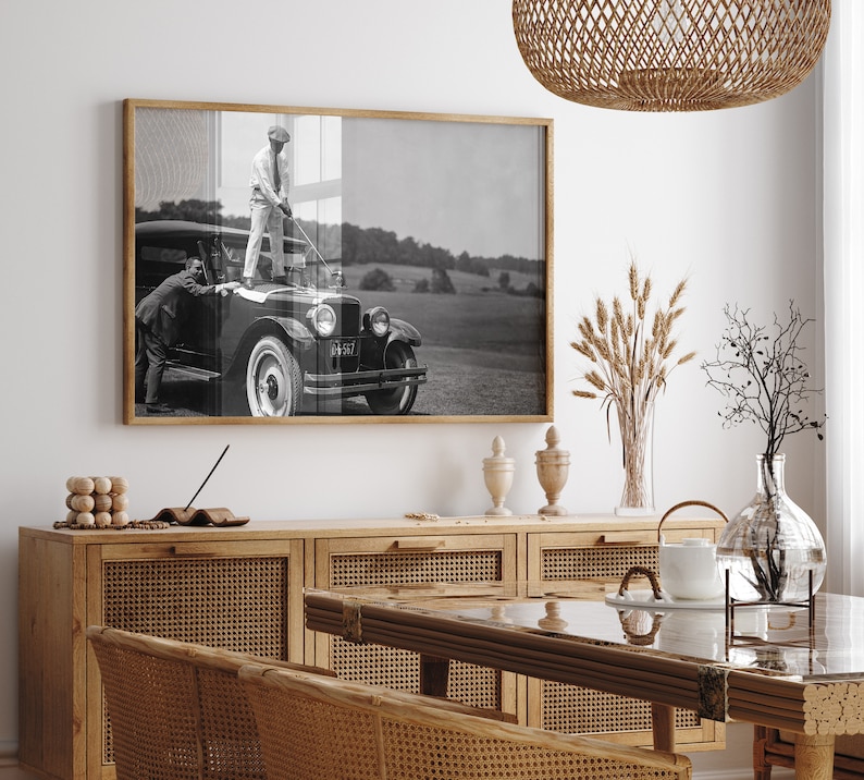 Op de afbeelding: Een zwart-witfoto van een man die op de treeplank van een vintage auto staat en een golfclub vasthoudt. De auto staat geparkeerd op een grasveld. De foto is ingelijst en hangt aan een witte muur boven een houten kast met rieten deuren.