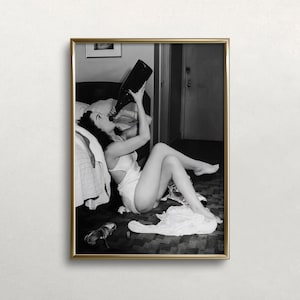 May include: A black and white photograph of a woman in a white slip dress drinking from a bottle of wine. She is sitting on the floor next to a bed with a white bedspread.