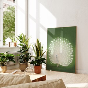 Op de afbeelding: Een ingelijste print van een witte pauw met zijn staartveren uitgespreid tegen een groene achtergrond. De print hangt aan een witte muur in een kamer met planten en een raam.