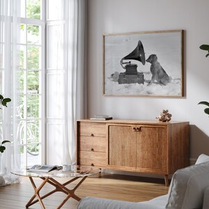 May include: A black and white photo of a dog listening to a vintage gramophone. The photo is framed and hanging on a white wall above a wooden cabinet with woven cane doors.