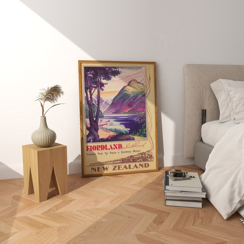 Puede incluir: Un p&oacute;ster de viaje vintage enmarcado para Fiordland, Nueva Zelanda, que presenta un paisaje esc&eacute;nico con monta&ntilde;as, un lago y un tren. El p&oacute;ster tiene un marco de madera y est&aacute; apoyado contra una pared blanca. Una peque&ntilde;a mesa de madera y un jarr&oacute;n est&aacute;n al lado del p&oacute;ster.
