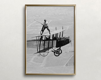 Op de afbeelding: Een zwart-witfoto van een man die op de vleugel van een vintage biplane in de lucht staat. De man houdt een stok in zijn hand en kijkt naar de camera. De biplane vliegt over een veld.