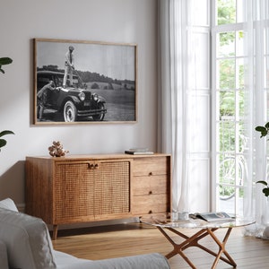 Op de afbeelding: Een zwarte vintage auto met een kast in rieten stijl op de achtergrond. De auto staat geparkeerd voor een witte muur en een raam met witte gordijnen. De auto heeft een zwart-witfoto van een man in golfkleding die op de treeplank van de auto staat. De foto is ingelijst in een zwart frame.