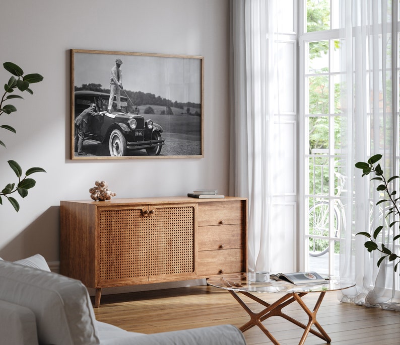 Op de afbeelding: Een zwarte vintage auto met een kast in rieten stijl op de achtergrond. De auto staat geparkeerd voor een witte muur en een raam met witte gordijnen. De auto heeft een zwart-witfoto van een man in golfkleding die op de treeplank van de auto staat. De foto is ingelijst in een zwart frame.