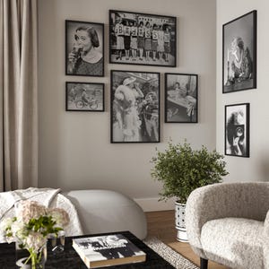 May include: A living room interior with a white sofa, a black coffee table, and a white armchair. The wall is decorated with black framed photographs in various sizes. The photographs are black and white and feature people and scenes from the past. A potted plant sits on the floor next to the armchair.